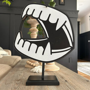 a laser cut graphic of Dracula's dentures hanging on a little wreath stand. in the background is a paneled wall with scallop trip painted black. it is ery cutsie