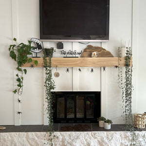 fireplace on a shiplap wall with a wood mantel. It is decorated with plants, a wood cut out of a mountain range, a laser cut spider garland and false dracula teeth on a stand