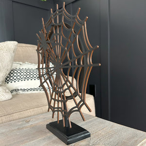 A wooden spider web hanging on a black stand. The background is a beige sofa and a wall of black paneling. 